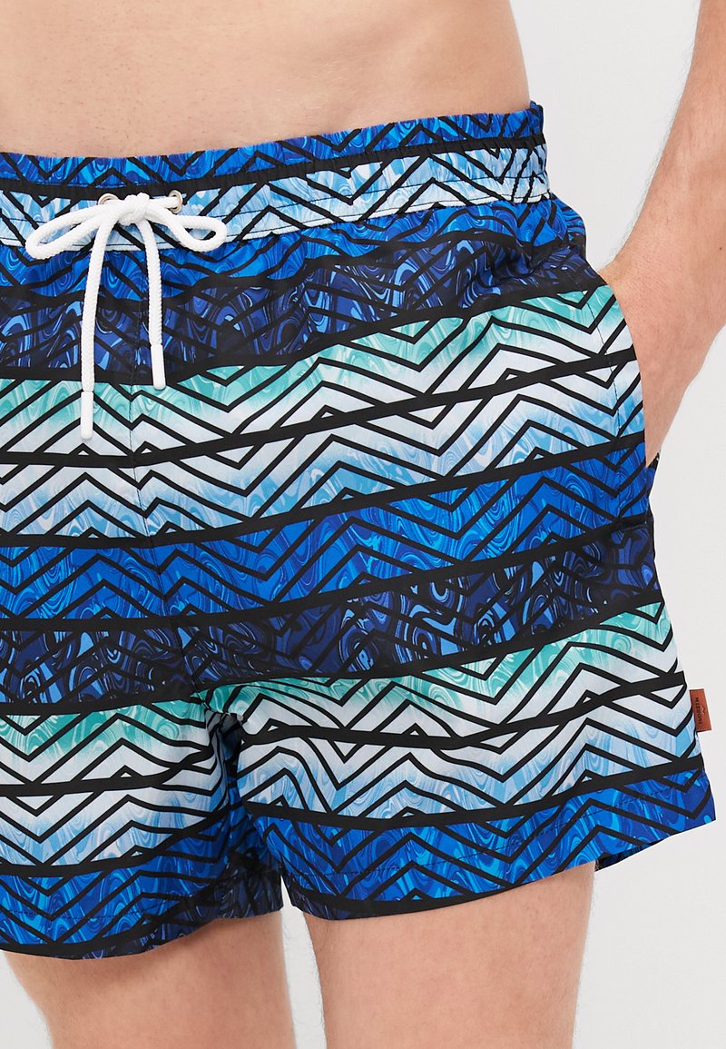 Blaue und türkisfarbene Badeshorts mit einem geometrischen Zickzackmuster und schwarzen Akzenten, ausgestattet mit einem Kordelzugbund und einer glatten Stofftextur.