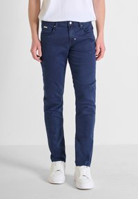 Tamno plave traperice od pamučnog materijala, slim-fit kroja, s pet džepova, prednjim kopčanjem na gumbe i diskretnim šavovima, uparenih s bijelim tenisicama.