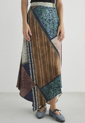 Frau trägt einen mehrfarbigen, asymmetrischen Wickelrock mit Blumen-, Streifen- und geometrischen Mustern, dazu graue Ballerinas.
