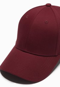 Mörkröd basebollkeps med en strukturerad krone, slät tyg, sex sydde ventilationshål och en böjd skärm. Har sömnadsdetaljer.