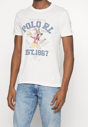 Biały t-shirt z kreskówkowym króliczkiem trzymającym marchewkę i monety, tekst "POLO RL EST. 1967" oraz mężczyzna w jasnoniebieskich dżinsach.