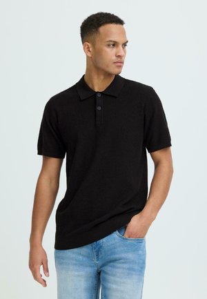 Jauns vīrietis, kas valkā melnu īsroku polokreklu un gaisā zilu džinsu bikses, skatās uz labo pusi, ar vienu roku kabatā.