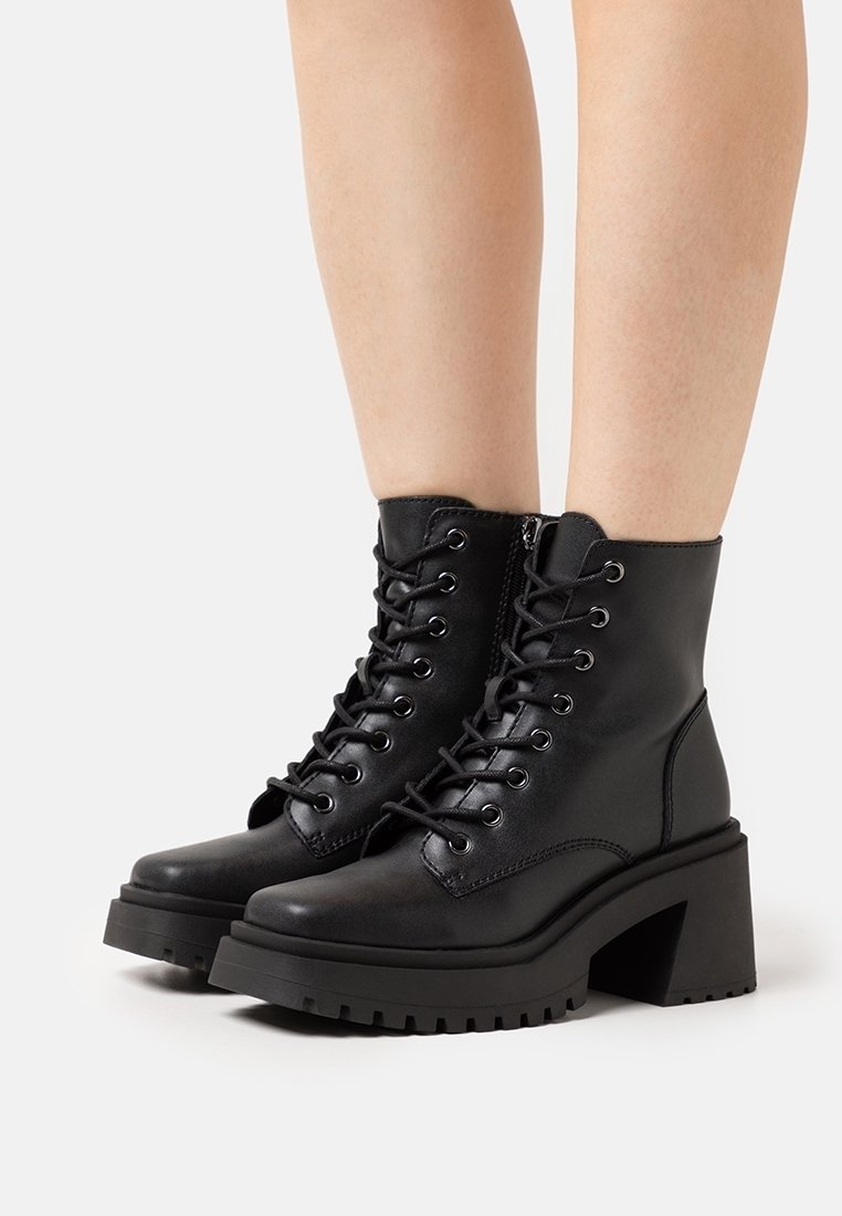 Bottines noires à lacets avec talons épais portées sur des jambes nues, présentées sur un fond blanc uni.