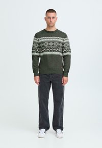 Pulover verde din lână tricotat, cu un model fair isle în nuanțe mai deschise, guler rotund, tiv cu nervuri, asortat cu blugi negri și adidași albi.