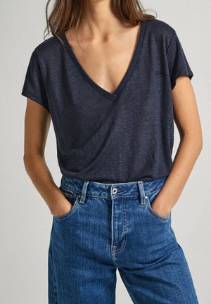 Woman wearing a dark navy V-neck short sleeve shirt tucked into high-waisted blue jeans with hands in pockets.