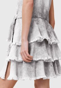 Vestido de mezclilla gris con diseño de volantes en capas y bordes deshilachados. La abertura lateral añade movimiento. Primer plano de la textura de la tela visible.