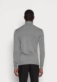 Homme portant un pull col montant gris ajusté et un pantalon noir, se tenant de dos contre un fond blanc uni.