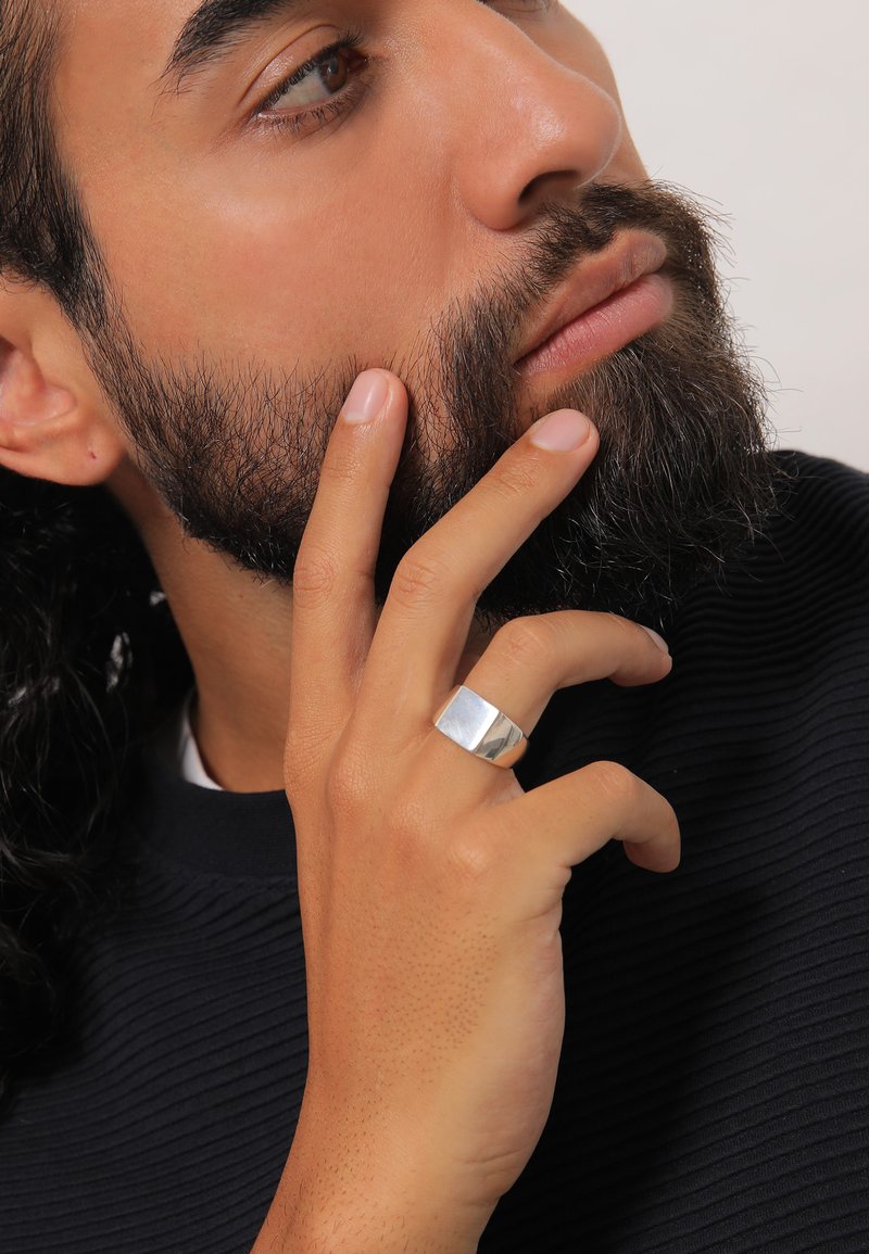 Silver ring with a flat, polished surface worn on a hand; textured fabric top in dark color; focus on hand and ring details.