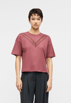 Młoda osoba o krótkich ciemnych włosach nosi luźną, pudrowo-różową koszulkę z koronkowym wycięciem w kształcie litery V oraz ciemne plisowane spodnie, stojąc na jasnym tle.