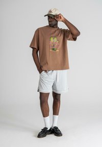 Brown graphic t-shirt with cactus design, light gray mesh shorts, black shoes, and a beige cap with text. Simple, relaxed fit, and casual style.