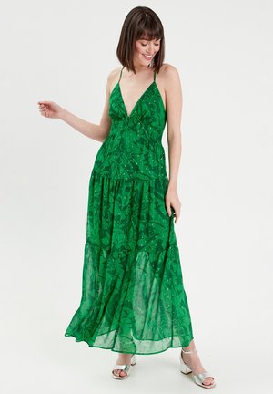 Robe maxi verte avec décolleté en V, bretelles spaghetti et ourlet à étages. Présente un motif de feuilles et une texture transparente. Associée à des sandales à talons argentées.