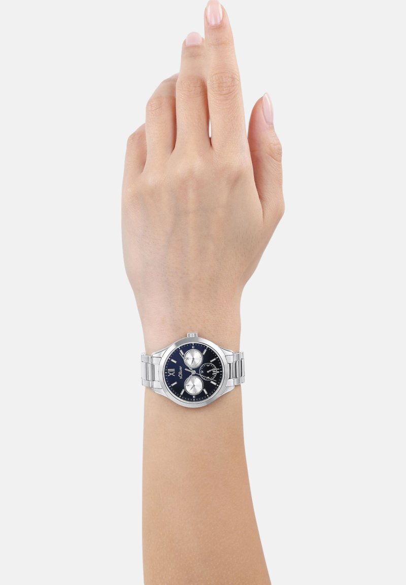 Silver metal watch on a wrist, featuring a dark blue dial, three subdials, Roman numerals, and a polished stainless steel band.