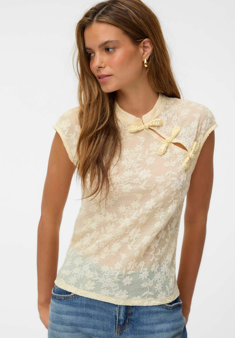 Femme portant un haut en dentelle florale crème transparente avec manches courtes et boutons décoratifs en forme de nœuds, associé à un jean en denim bleu.