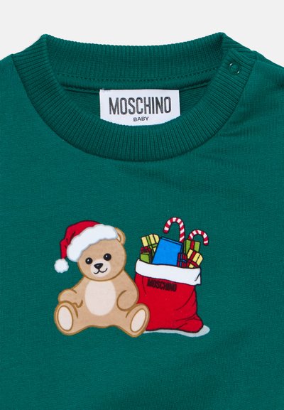 Πράσινο μπλουζάκι μωρού με ετικέτα Moschino, με ένα αρκουδάκι που φοράει καπέλο Άγιου Βασίλη δίπλα σε μια κόκκινη σακούλα γεμάτη δώρα και καραμέλες.
