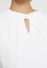 Bílá blůza s výstřihem ve tvaru klíčové dírky, plisovaným designem a hladkou texturou. Tkanina vypadá lehce a jemně obepíná ramena.