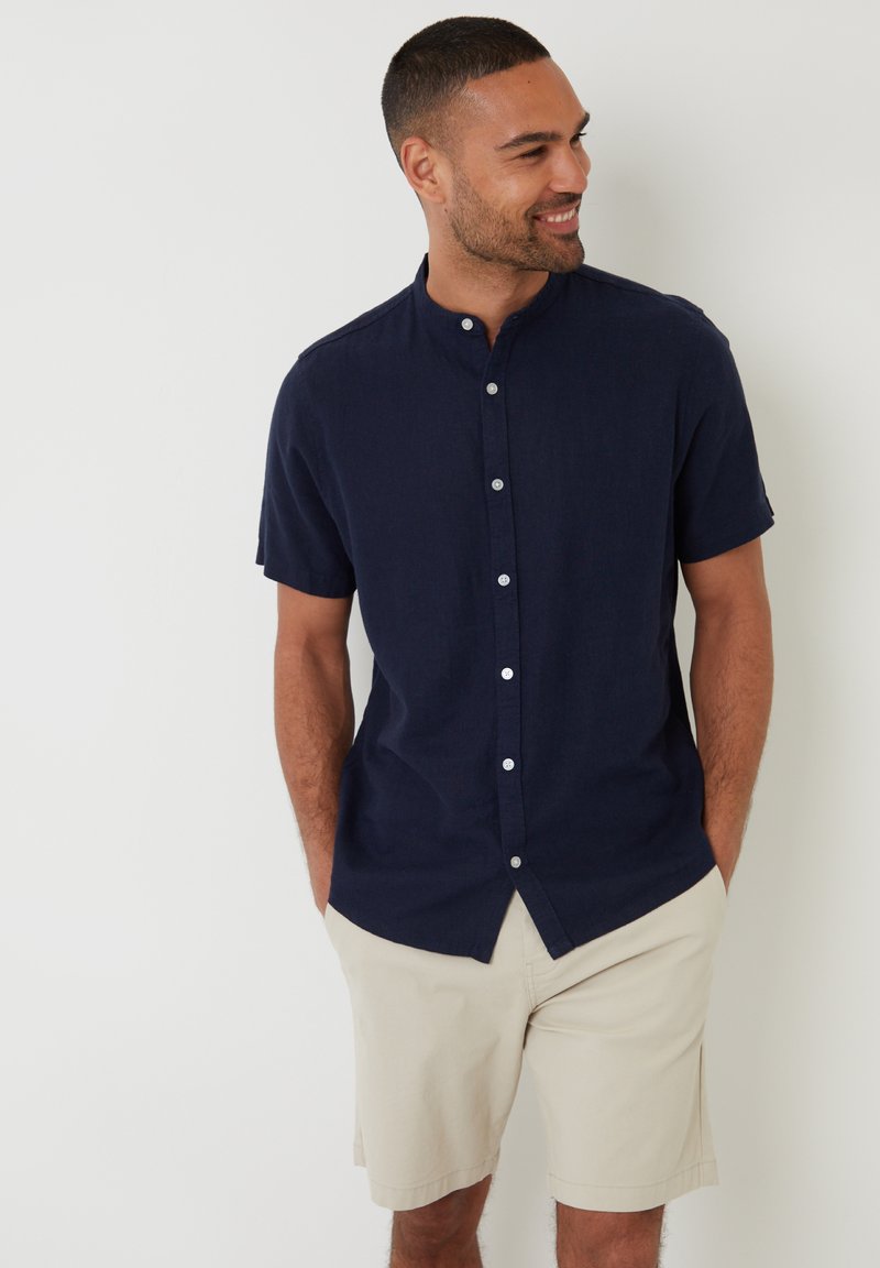 Marineblauw shirt met korte mouwen en een knoopsluiting aan de voorkant, gemaakt van een lichtgewicht stof. Gecombineerd met khaki shorts, handen in de zakken, casual staand.