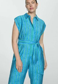 Donna che indossa un tuta senza maniche a righe verticali blu e verde, con una cintura annodata e orecchini a cerchio in argento, su uno sfondo semplice.