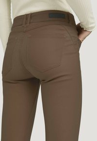 Pantalones ajustados marrones de una tela suave y elástica, con bolsillos traseros y una etiqueta negra en la cintura, que enfatizan una silueta ajustada.