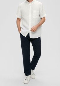 Man in een witte overhemd met korte mouwen, donkere marineblauwe broek en witte sneakers, staande met één hand in zijn zak.