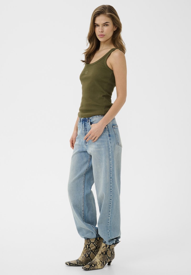 Femme portant un débardeur vert olive, un jean bleu clair ample et des bottines pointues à motif serpent, debout devant un fond uni.