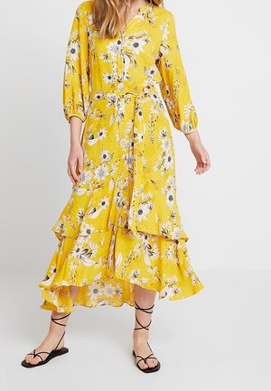 Kvinde iført en gul blomstret midi-knapkjole med flæsekant og matchende bælte, kombineret med sorte flade sandaler med stropper.