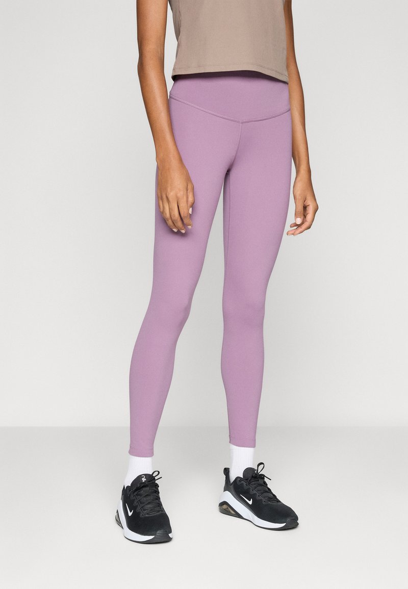 Persona vestida con leggings de tiro alto en color lila, calcetas blancas y zapatillas Nike negras, de pie frente a un fondo claro y liso.