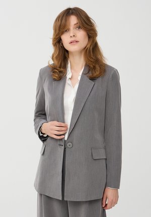 Blazer grigio su misura con risvolti, chiusura con un bottone, due tasche frontali e una texture liscia, abbinato a una camicia bianca.