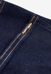 Dark blue denim fabric with double beige stitching and a close-up of a copper rivet reinforcing a pocket corner.