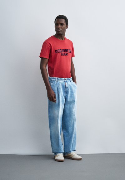 Άνδρας που φοράει κόκκινο μπλουζάκι με σχέδιο, φαρδιά ανοιχτό μπλε τζιν και λευκά αθλητικά παπούτσια, στέκεται μπροστά σε μια μονόχρωμη αχνή τοίχο.