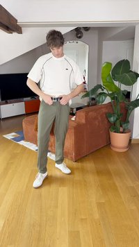 Fehér rövidujjú ing fekete részletekkel, olivazöld nadrág és fehér sportcipő. Egy nappaliban áll, ahol fa padló van és egy növény.