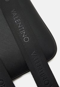 Juodas tekstūruotas maišelis su lygiu paviršiumi ir išryškintu embosiuotu "VALENTINO" logotipu ant storos dirželio. Minimalistinis dizainas su švariomis linijomis.