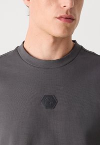 Gray short-sleeve shirt with a textured fabric. Features a hexagonal black logo on the chest and a round neck design.