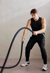 Zwarte battle ropes vastgehouden door een persoon in een mouwloos zwart topje en zwarte broek. Witte sportschoenen met rode accenten. Glad, licht achtergrond.