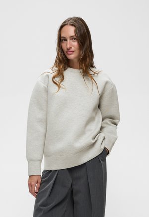 Femme aux longs cheveux bruns portant un sweat-shirt gris clair oversized et un pantalon plissé gris foncé, debout devant un fond uni.