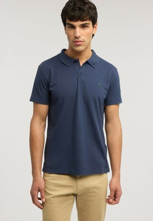 Giovane uomo con polo a maniche corte blu navy e pantaloni beige, in piedi davanti a uno sfondo chiaro e uniforme.