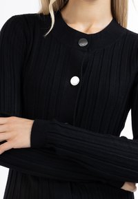 Kvinna som bär en svart ribbad långärmad kofta med stora silverknappar, med armarna korsade vid midjan, mot en vit bakgrund.