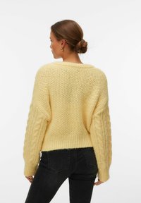Femme aux cheveux attachés en chignon, portant des boucles d'oreilles en or en forme de créoles, un pull en maille texturée jaune pâle, et un jean noir ajusté, debout de dos à la caméra.