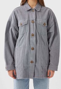 Vrouw in een grijze corduroy button-up jas met klepborstzakken en lichtblauwe jeans tegen een effen achtergrond.