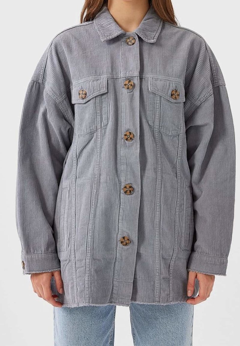 Vrouw in een grijze corduroy button-up jas met klepborstzakken en lichtblauwe jeans tegen een effen achtergrond.