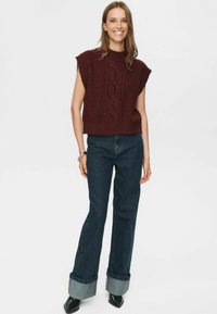 Besidinti moteris, dėvinti be rankovių bordo spalvos megztinį su pynėmis, tamsius, pasukamus džinsus, juodus smailius batus, ir ant dešinės rankos nešiojanti apyrankes.