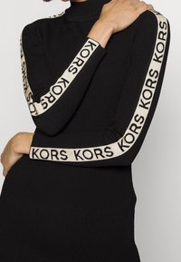 Donna che indossa un vestito lungo a maniche lunghe nero a costine con strisce bianche con il logo "KORS" che scorrono lungo le braccia e il torso, braccia incrociate.