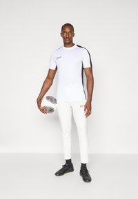T-shirt sporty blanc Nike avec des accents noirs, associé à un pantalon slim blanc avec des rayures roses, tenu avec un ballon de soccer à motifs.