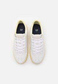 Sneaker bianche in pelle con suole e dettagli gialli, caratterizzate da un design testurizzato, lacci rotondi e il logo "GAP" sulla linguetta.