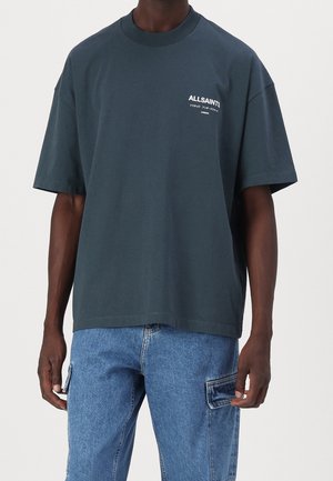 Mand iført mørkegrå ALLSAINTS logo t-shirt og blå denim cargo bukser, stående mod en ensartet lys baggrund.