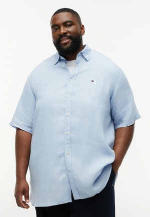 Lächelnder Mann mit Bart, der ein hellblaues kurzärmeliges Hemd mit Knöpfen und eine dunkle Hose trägt, stehend mit einer Hand in der Tasche.