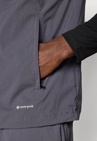 Gray, lightweight jacket made of nylon, featuring a side pocket with a drawstring, and a logo embroidered near the hem. Black long-sleeve shirt underneath.