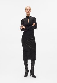 Vestido preto de manga longa com colarinho alto e detalhe em forma de chave na frente, feito de tecido texturizado com um brilho sutil, combinado com botas pretas até ao joelho.