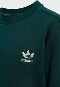 Sudadera verde oscuro hecha de una tela suave, con cuello redondo y un logo de Adidas bordado en blanco con diseño de trébol.