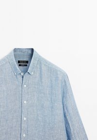 Light blue button-up shirt made of textured fabric. Features a classic collar, six buttons, and a label visible under the collar.