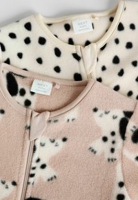 Dwa dziecięce ubrania z polarowego materiału, jedno kremowe w czarne kropki, a drugie w odcieniu różowym z czarnymi i kremowymi wzorami. Oba mają okrągłe dekolty i zamki błyskawiczne.